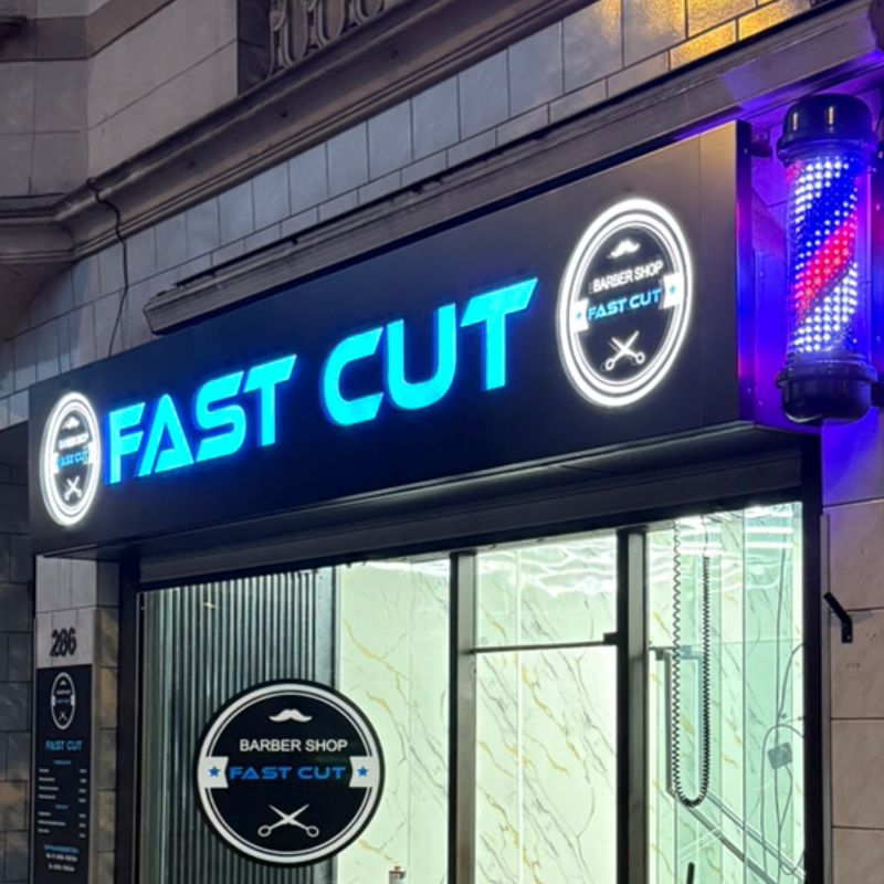Das Schild von Fast Cut am Abend: blau leuchtende Buchstaben, zwei weiße Leuchtplaketten und der Barber-Pole