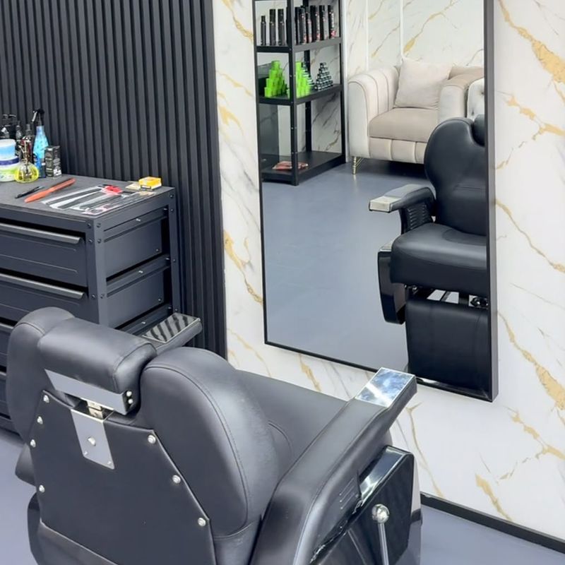 Schwarzer Barberstuhl mit Chromdetails vor einem Standspiegel, dahinter weiße Marmor-Optik mit Goldadern und schwarze Lamellen